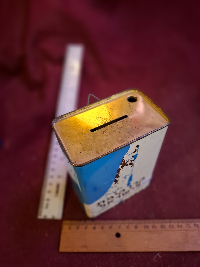 Vintage metal coin bank with slot, distressed paint, placed beside measuring rulers