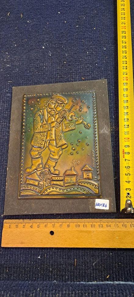 Vintage embossed metal plaque with fiddler and village scene, rulers for scale, collectible art