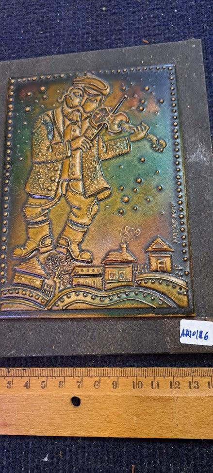 Embossed metal art of a man playing violin above rustic houses, vintage collectible decor