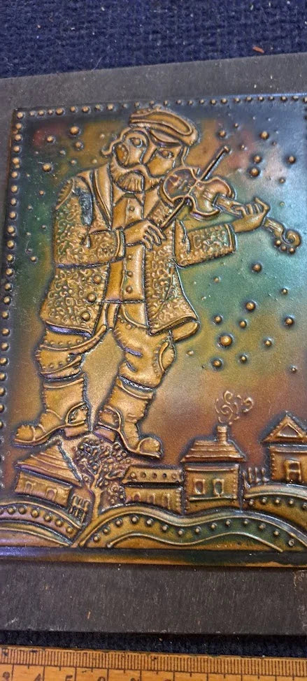 Vintage metal embossing of a man playing violin above village houses, rustic collectible decor.