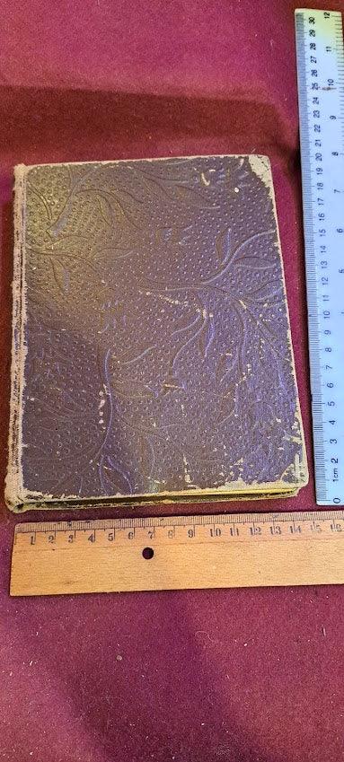 Vintage embossed hardcover book with worn edges on a maroon surface, two rulers for scale