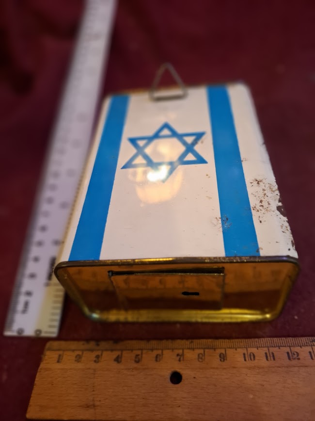 Vintage Israeli tin charity box with blue Star of David, measured with rulers, on red fabric.