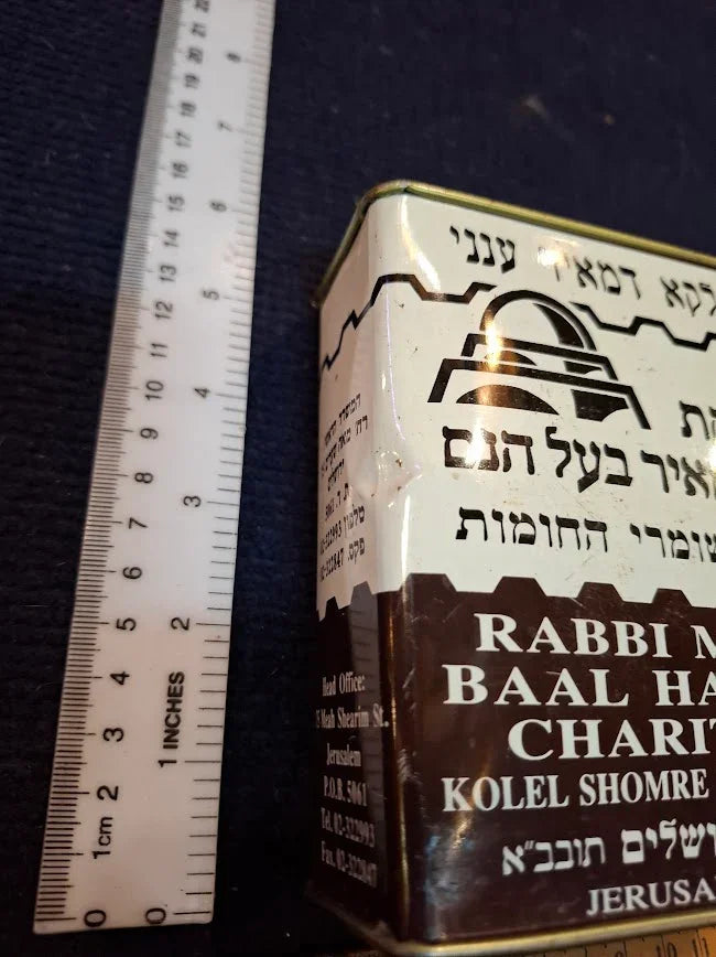 Vintage metal charity box with Hebrew text beside ruler for size reference, collectible item