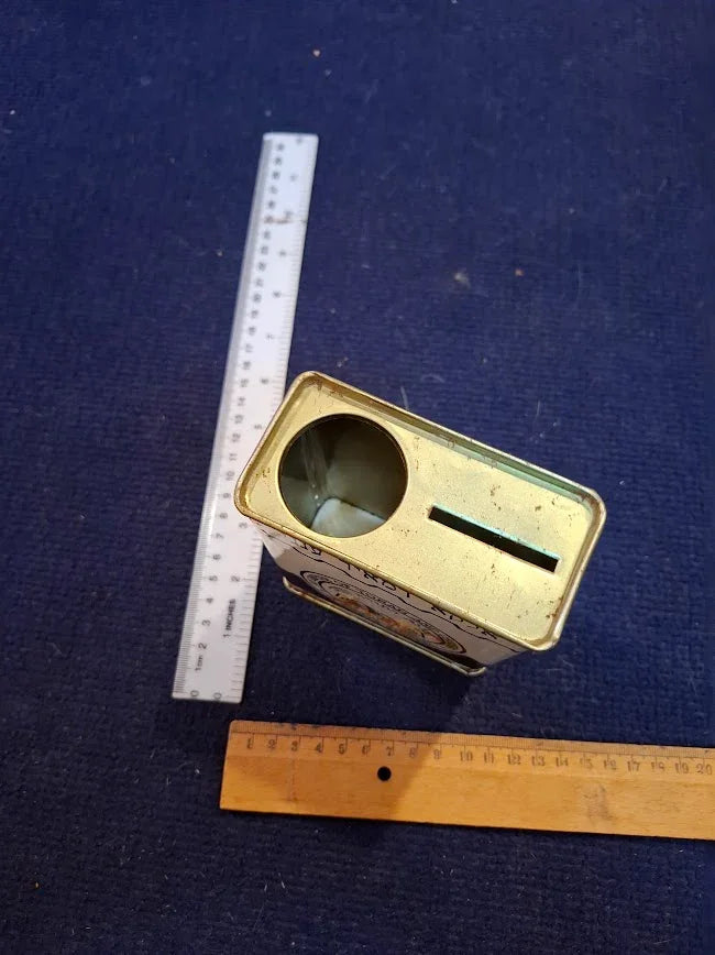 Vintage metal coin bank tin with slot and circular opening, rulers for scale, on blue fabric