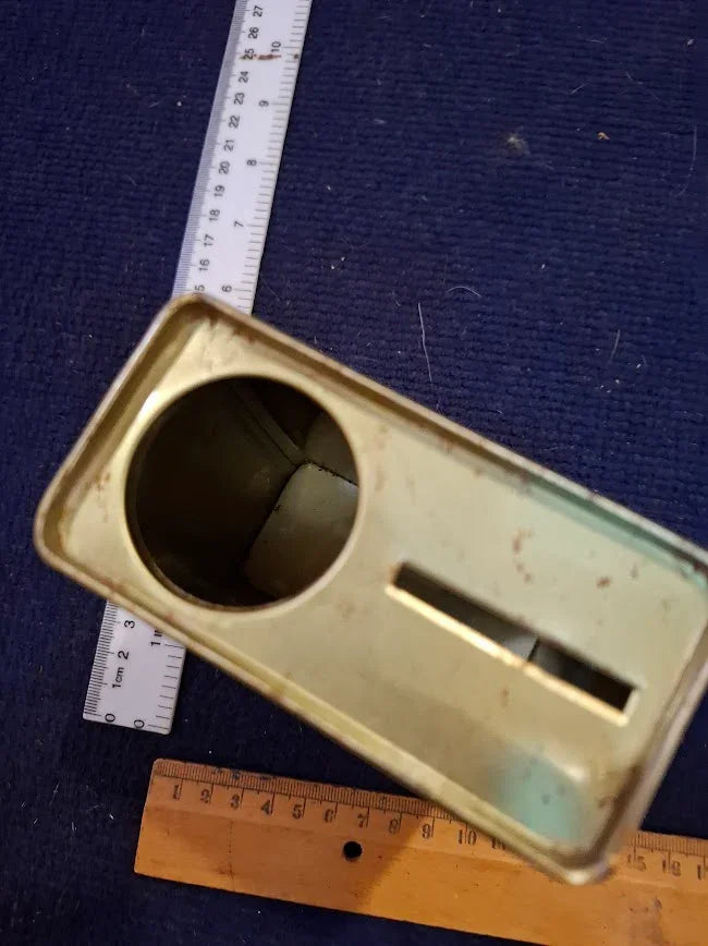 Vintage metal tin with coin slot, circular opening, and rulers for scale on blue fabric