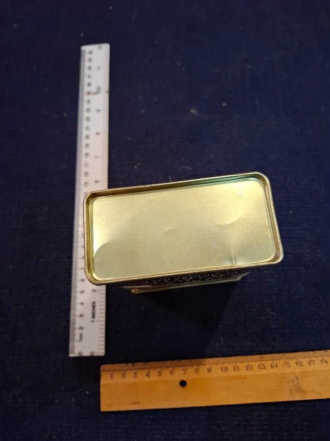 Rectangular vintage metal tin with gold base, shown with rulers for size reference on dark surface