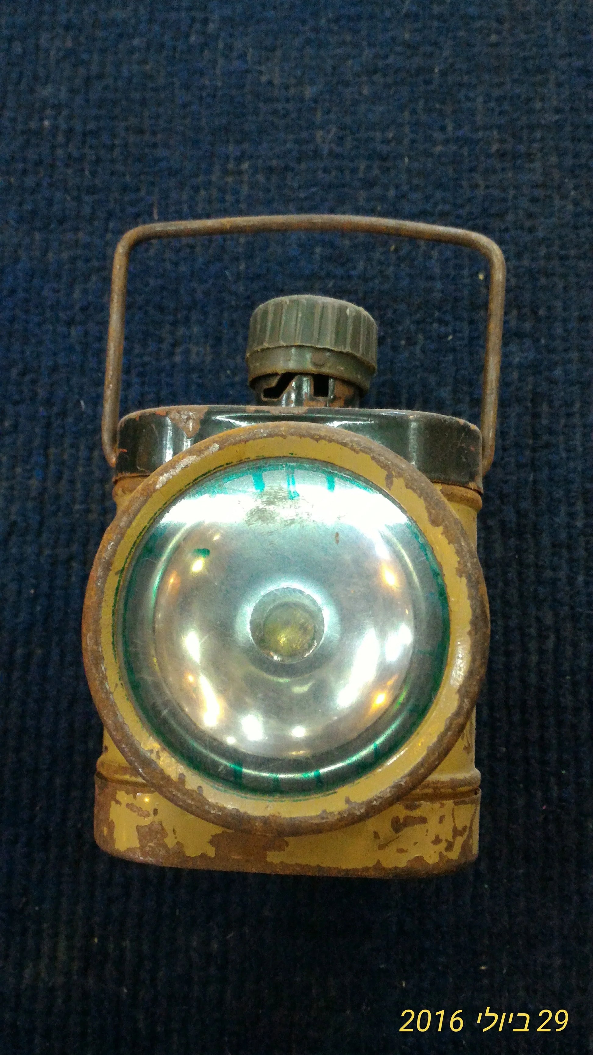 Vintage metal flashlight with handle and round lens, showing rust and wear on blue fabric